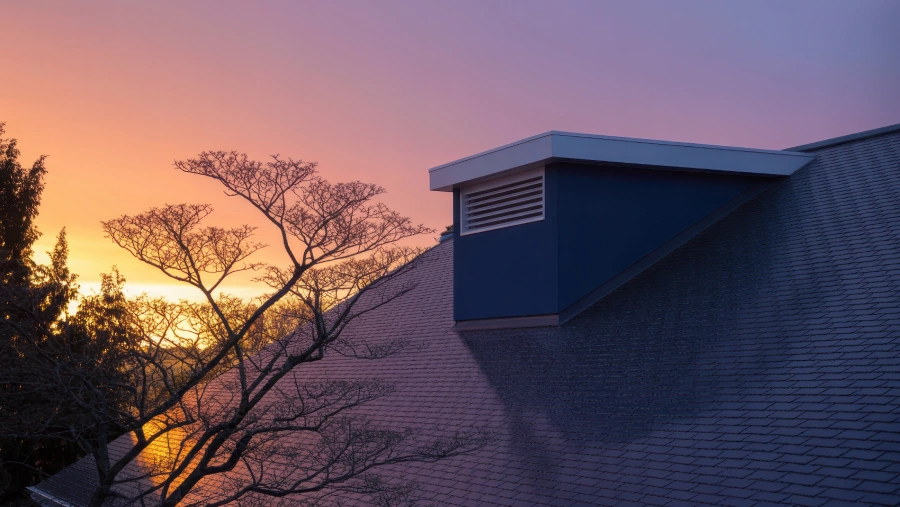 Storm Damage Restoration services Modern house roof with dark grey shingles under a dramatic sunset sky and tree silhouette